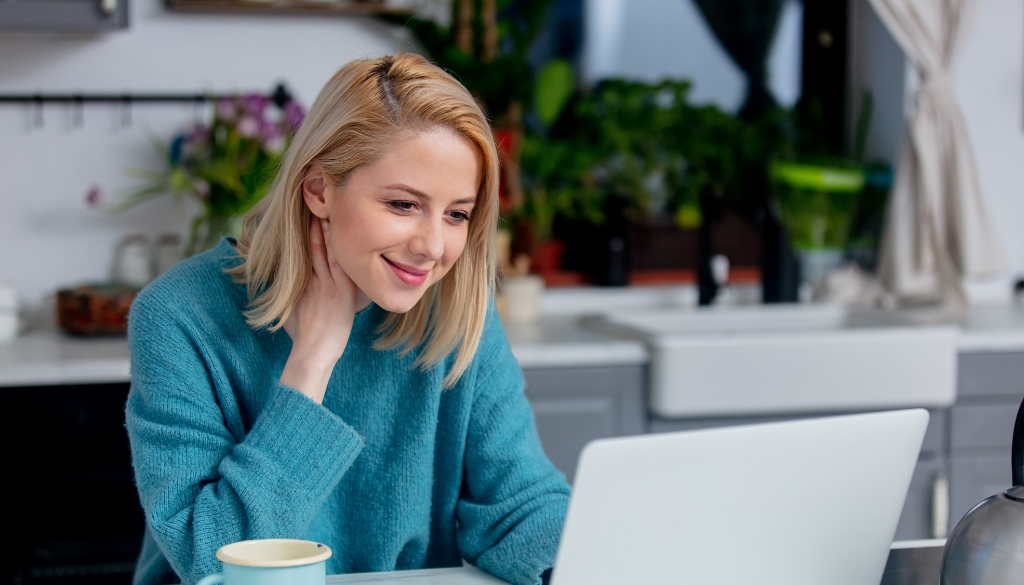 Woman taking a call on her laptop while working from home during the COVID-19 Pandemic.|Woman smiling while working remotely taking a video call on her laptop.|Woman smiling while on a video call during the COVID pandemic.